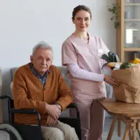 An elderly man sitting on a wheelchair and a female caregiver preparing a meal