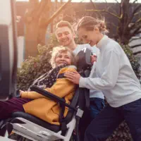 An elderly woman sitting on a wheelchair and being assisted by two staff to get her in to the van